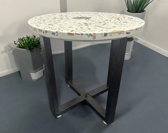 Pode incluir: Uma mesa auxiliar redonda com tampo de terrazzo branco e estrutura de metal preto. A mesa tem um design simples e moderno e seria uma ótima adição a qualquer sala de estar ou quarto.