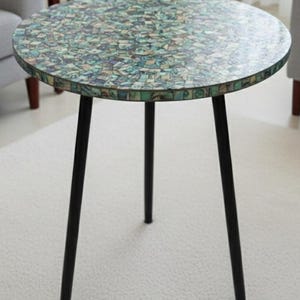 Könnte beinhalten: Runder Beistelltisch mit einem Mosaik-Aufsatz in Grün-, Blau- und Beigetönen. Der Tisch hat drei schwarze Metallbeine. Der Tisch hat einen Durchmesser von ca. 61 cm und ist 51 cm hoch.