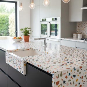 Könnte beinhalten: Moderne Kücheninsel mit einer weißen Arbeitsplatte mit einem farbenfrohen Terrazzo-Muster. Die Insel hat ein weißes Bauernhaus-Waschbecken und schwarze Schränke. Überkopf beleuchten drei Pendelleuchten den Raum. Ein Wasserhahn aus Edelstahl ist installiert.