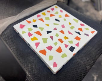 Pode incluir: Azulejo quadrado branco com um design de terrazzo colorido. O azulejo apresenta formas irregulares em rosa, laranja, amarelo, verde e preto. O azulejo tem aproximadamente 25 cm quadrados.