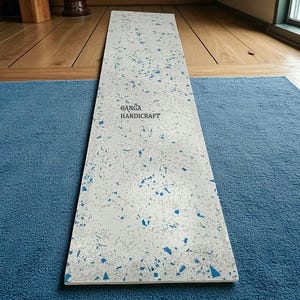 May include: A rectangular, white decorative board with blue speckles and the text "GANGA HANDICRAFT" in the upper centre. The board is placed on a blue rug, with a wooden floor in the background.