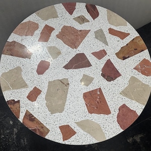 Puede incluir: Una tapa de mesa redonda con fondo blanco y diseño de terrazo. La tapa de mesa está hecha de pequeños trozos de mármol rojo, marrón y blanco.