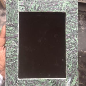 Könnte beinhalten: Rechteckiger Bilderrahmen mit grünem und lila Marmormuster. Der Rahmen hat einen schwarzen Innenraum und besteht aus einem steinartigen Material. Der Rahmen ist für ein Foto oder Kunstwerk konzipiert.