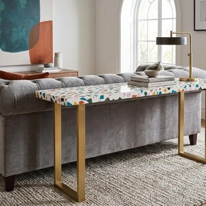 May include: A rectangular console table with a terrazzo top featuring colorful chips. The table has gold-colored metal legs and is positioned in front of a gray upholstered sofa. A black lamp and books sit on the table.