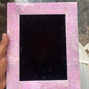 Può includere: Una cornice rettangolare con un bordo rosa simile a pietra screpolata. L'interno della cornice è nero, creando un contrasto con l'esterno rosa chiaro. La cornice è progettata per contenere una fotografia o un'opera d'arte.