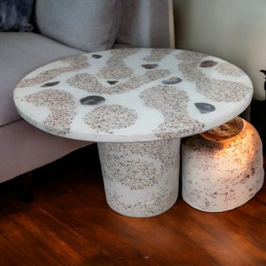 May include: A white round side table with a unique speckled finish. The table top has a pattern of white and gray swirls with embedded stones. The table has a round base with a smaller round base underneath.