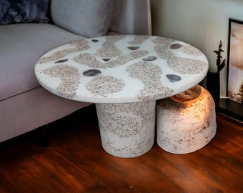 Pode incluir: Uma mesa auxiliar redonda branca com um acabamento único salpicado. O tampo da mesa tem um padrão de redemoinhos brancos e cinzentos com pedras incrustadas. A mesa tem uma base redonda com uma base redonda menor por baixo.