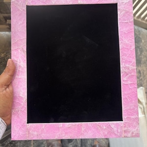 Può includere: Una cornice rettangolare rosa con interno nero. La cornice ha un aspetto strutturato, simile a un mosaico, con sfumature di rosa e bianco. La cornice è tenuta da una persona.