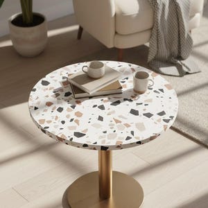 May include: A circular side table with a terrazzo-style surface in white, black, beige, and brown. The table features a gold-coloured cylindrical leg and base. Two mugs and books are on the table.