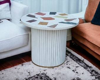 Pode incluir: Uma mesa auxiliar redonda branca com uma base branca e um tampo de terrazzo com manchas coloridas. A mesa está sobre um tapete com fundo branco e padrões pretos e cinzentos.