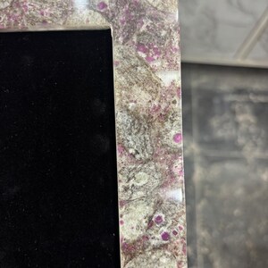 Könnte beinhalten: Ein rechteckiger Rahmen mit einer polierten Oberfläche aus einem Mosaik aus grauen, weißen und beigen Steinfragmenten, durchsetzt mit leuchtend magentafarbenen Mineraleinschlüssen, der eine tiefschwarze, samtartige Innenfläche umrandet.