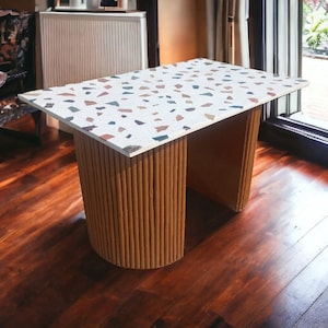 May include: A small, rectangular table with a white terrazzo tabletop and a brown cardboard base. The table is in a room with hardwood floors and a window in the background.