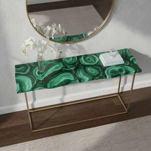 Puede incluir: Una consola rectangular con una tapa con patrón de malaquita y patas de metal dorado. La mesa está decorada con un jarrón de cristal transparente con flores blancas, una pequeña pila de libros y una esfera decorativa de cristal. La mesa mide 122 cm de largo.