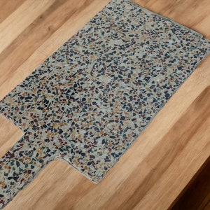 May include: A rectangular terrazzo cutting board with a handle. The board is made of a light gray base with brown, black, and red flecks.
