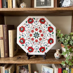 May include: An octagonal white marble plaque featuring intricate pietra dura inlay work. The design displays a symmetrical floral pattern with vibrant red, green, and blue stone petals arranged in a circular motif, supported by a black stand.