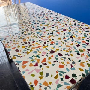 Könnte beinhalten: Ein rechteckiger Tisch mit einer weißen Oberfläche, die mit einem farbenfrohen Terrazzo-Muster verziert ist. Das Design zeigt ein Mosaik aus unregelmäßigen Formen in Orange-, Blau-, Grün- und Burgundertönen. Die Tischbeine sind sichtbar, und die Oberfläche reflektiert Licht.