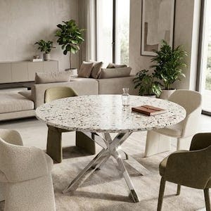Op de afbeelding: Een ronde eettafel met een wit terrazzo blad met veelkleurige steensplinters en een gepolijst verchroomd metalen onderstel met kruisende poten. De tafel is omringd door vier gestoffeerde fauteuils in crème en olijfgroene stof.