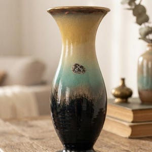 Może przedstawiać: Ceramiczny wazon z rozszerzanym brzegiem i zwężaną szyjką, posiadający gradientowe szkliwo przechodzące od złocistożółtego u góry, przez turkusowy w środku, aż po głęboką czerń u podstawy. Owalny emblemat ze skrzyżowanymi kluczami znajduje się na szyjce.
