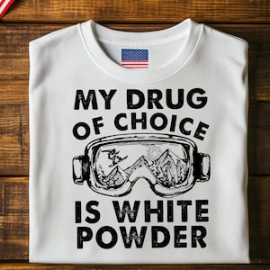 May include: White t-shirt with the text "MY DRUG OF CHOICE IS WHITE POWDER" in black. The design includes a graphic of ski goggles with a skier and mountain scene. An American flag patch is visible.