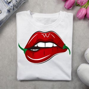 Può includere: T-shirt bianco con grafica di labbra rosse che mordono un peperoncino verde. Le labbra sono di un rosso vibrante con denti bianchi. La maglietta è piegata su una superficie grigia chiara, con un paio di jeans e scarpe da ginnastica.