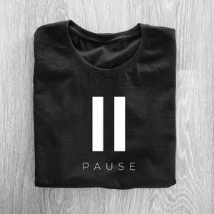 May include: Black t-shirt with a white pause symbol, two vertical bars, and the word "PAUSE" printed below. The shirt is folded neatly on a light wood-grain surface.