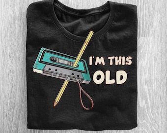 May include: Black t-shirt featuring a teal cassette tape with a pencil through it and the text "I'M THIS OLD" in white and beige. The shirt is folded and has a crew neck.
