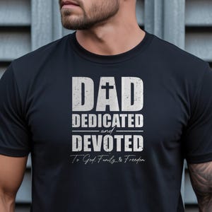 Peut inclure: Un t-shirt noir à manches courtes avec un texte blanc vieilli indiquant DAD avec une croix dans la lettre A, suivi de DEDICATED et DEVOTED, et le texte manuscrit To God, Family & Freedom centré sous le motif graphique principal.