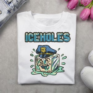 Peut inclure: T-shirt blanc avec le texte "ICEHOLES" en bleu et un dessin animé d'un glaçon portant un chapeau de policier. Le glaçon a un visage et est entouré de gouttelettes d'eau. Le t-shirt est sur une surface grise.