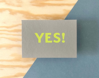 Tarjeta postal serigrafiada con un diseño tipográfico sencillo, tarjeta con el eslogan "¡Sí!", impresa a mano, con el logotipo estampado en el reverso.