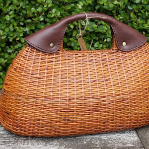 Könnte beinhalten: Eine braune Korbtasche mit einem braunen Ledergriff und einer braunen Lederklappe. Die Tasche hat eine geflochtene Textur und eine abgerundete Form.