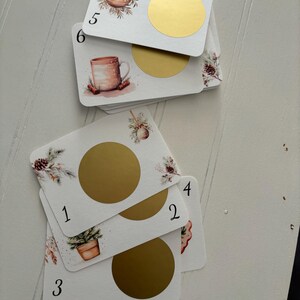 Peut inclure: Plusieurs cartes rectangulaires blanches avec des cercles dorés à gratter. Chaque carte présente une illustration à l'aquarelle d'objets sur le thème des fêtes, tels que des ornements, une tasse et une plante en pot. Les numéros 1 à 6 sont imprimés.