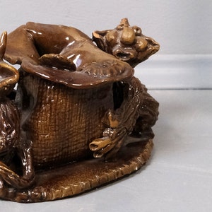 Può includere: Una statuetta in ceramica di due creature mitiche marroni, una seduta in un vaso marrone, l'altra appollaiata sul bordo. Le creature hanno grandi orecchie e musi lunghi.