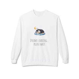 Puede incluir: Sudadera blanca con un pingüino de dibujos animados durmiendo sobre un témpano de hielo bajo una luna creciente. El texto "DREAMS LOADING... PLEASE WAIT." está impreso debajo. La sudadera tiene mangas largas y cuello redondo.