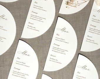 May include: White, semi-circular menu cards with elegant script text, arranged on a neutral-toned surface. The cards feature menu items like Couscous Royal and Wedding Cake. Dried flowers add a decorative touch.