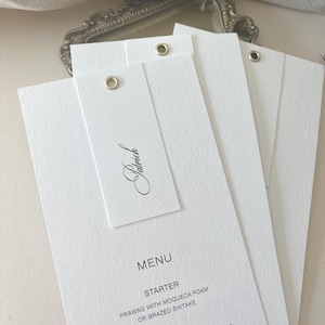 May include: Three white rectangular menu cards with textured paper, each featuring a gold metal eyelet at the top. The top card displays the name Patrick in black script, followed by a menu listing starters and main courses in black text.