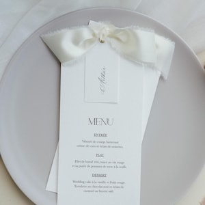 Peut inclure: Une carte de menu en papier blanc avec du texte noir indiquant MENU, suivi des sections ENTREE, PLAT et DESSERT. Une petite étiquette blanche portant le nom Arthur est fixée en haut avec un ruban de soie crème aux bords effilochés.