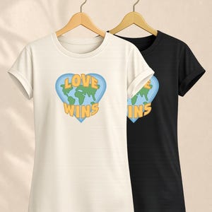 Könnte beinhalten: Ein cremefarbenes Kurzarm-T-Shirt und ein schwarzes Kurzarm-T-Shirt, beide mit einer Grafik eines hellblauen Herzens, das eine grüne Weltkarte und den gelben Schriftzug LOVE WINS enthält, präsentiert auf Holzkleiderbügeln.