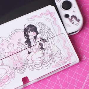 Puede incluir: Consola Nintendo Switch blanca con una piel personalizada que presenta un ángel rosa y blanco con alas, un corazón y una cruz. La piel también incluye una cinta rosa y plumas.