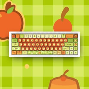 Op de afbeelding: Een mechanisch toetsenbord met een aangepaste set toetsen met een fruit- en dessertthema. De toetsen zijn gekleurd in tinten oranje, groen en crème en tonen diverse iconen zoals appels, kersen en pudding, naast de standaard letters.