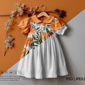 Könnte beinhalten: Ein weißes Kleid mit einem Blumendruck auf dem Oberteil. Das Kleid hat einen Peter-Pan-Kragen und kurze Ärmel. Das Kleid hängt an einem Holzbügel vor einem braunen und grauen Hintergrund.