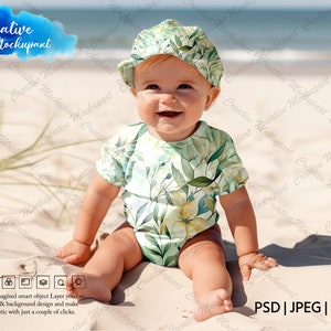 May include: A baby wearing a green and yellow floral print onesie and a green and white floral print hat sits on a sandy beach. The baby is smiling and looking at the camera.