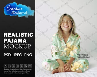 May include: A girl wearing a green and yellow floral pajama set sits on a white bed. She is smiling and has her hands in her lap.