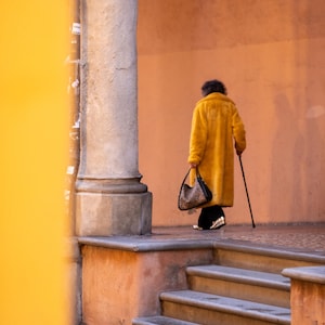 May include: A person wearing a yellow coat walks down a set of stairs with a handbag and a cane.
