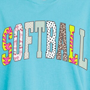 May include: A turquoise blue t-shirt with the word "SOFTBALL" in a multi-patterned design. The letters feature patterns like cherries, polka dots, and animal print. The shirt has a relaxed fit and short sleeves.