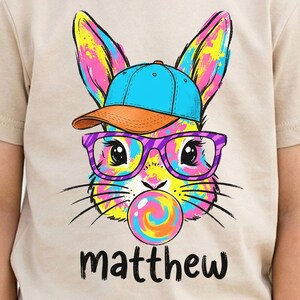 May include: Beige t-shirt featuring a colorful cartoon bunny wearing a blue cap, orange visor, and purple glasses, blowing a bubblegum bubble. The name "matthew" is printed in black script below the bunny.