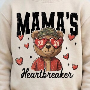 Puede incluir: Sudadera beige con un gráfico de un oso de peluche con gafas de sol en forma de corazón con "XO" en las lentes. El oso también lleva un gorro de camuflaje y una chaqueta roja. El texto "MAMA'S Heartbreaker" está impreso encima y debajo del oso.