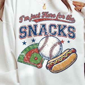 May include: White sweatshirt with a baseball-themed graphic. The design includes a baseball, a hot dog, and a baseball field, with the text "I'm just Here for the SNACKS" in red and blue lettering. Stars are also included in the design.