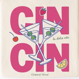 Puede incluir: Sudadera color crema con un gráfico rosa que dice "GIN" y "CIN". El diseño incluye dos copas de martini con aceitunas, una fresa, una cereza y un limón. El texto dice "la dolce vita" y "Cheers! Viva!"