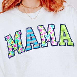 May include: White sweatshirt with the word "MAMA" in colourful, decorative lettering. The letters feature floral, striped, and gingham patterns with a scalloped navy blue border. A gold necklace is visible.