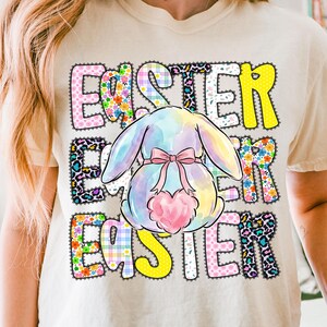 May include: A beige t-shirt with the word "EASTER" in colorful, patterned letters. A pastel-colored bunny with a pink bow and heart-shaped tail is centered on the shirt. The design has floral, leopard, and plaid patterns.